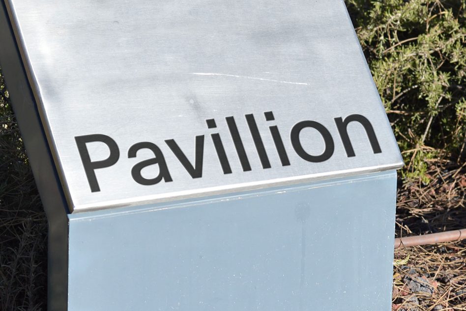 Light blue directional sign mounted on landscape post, reads Pavillion in bold sans serif