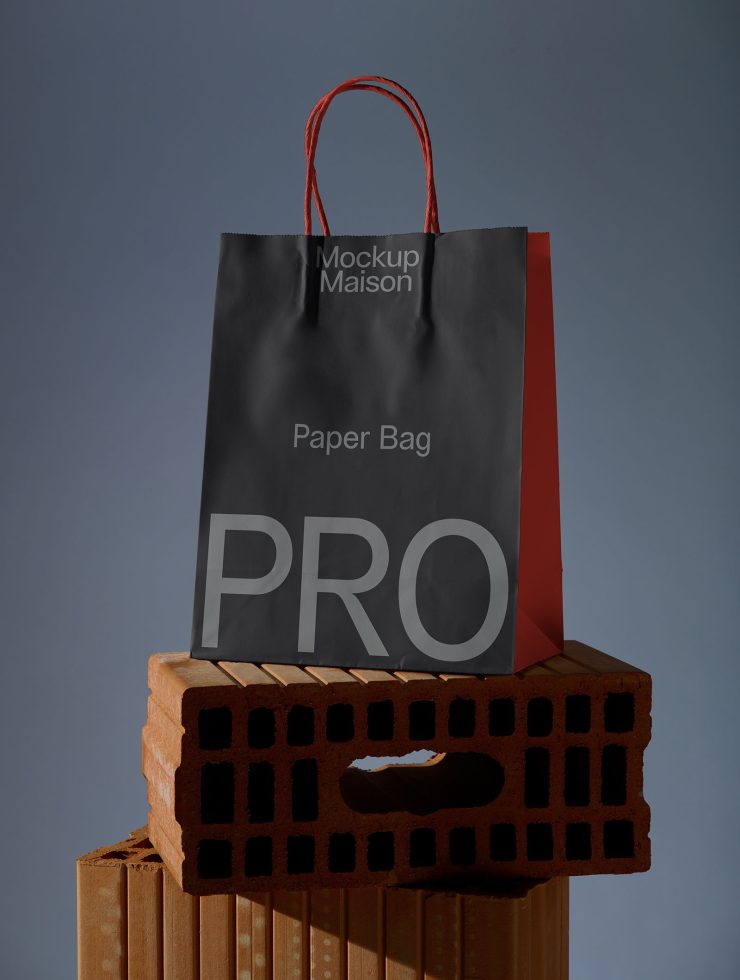 Black paper bag mockup with red handles on a brick stand against a blue background, ideal for design presentations and ecommerce.