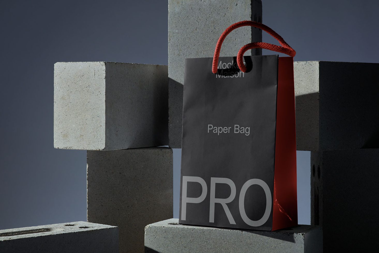 Black paper bag mockup with red rope handle set against concrete blocks for product design presentation, ideal for modern branding.