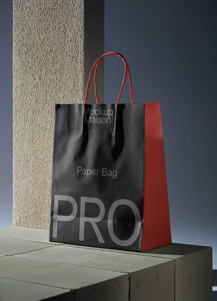 Elegant paper bag mockup with red handles on a concrete ledge for branding presentation, realistic shadow overlay, suitable for design templates.