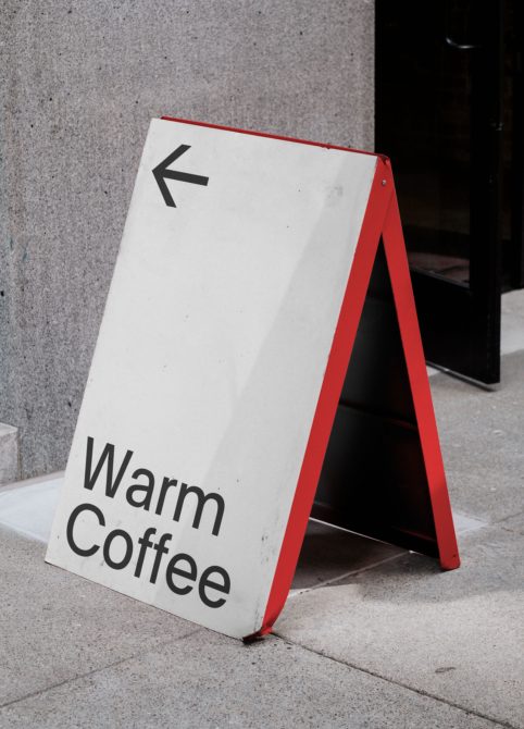 A-frame sidewalk sign mockup with Warm Coffee text and arrow, urban setting for outdoor advertising and branding design.