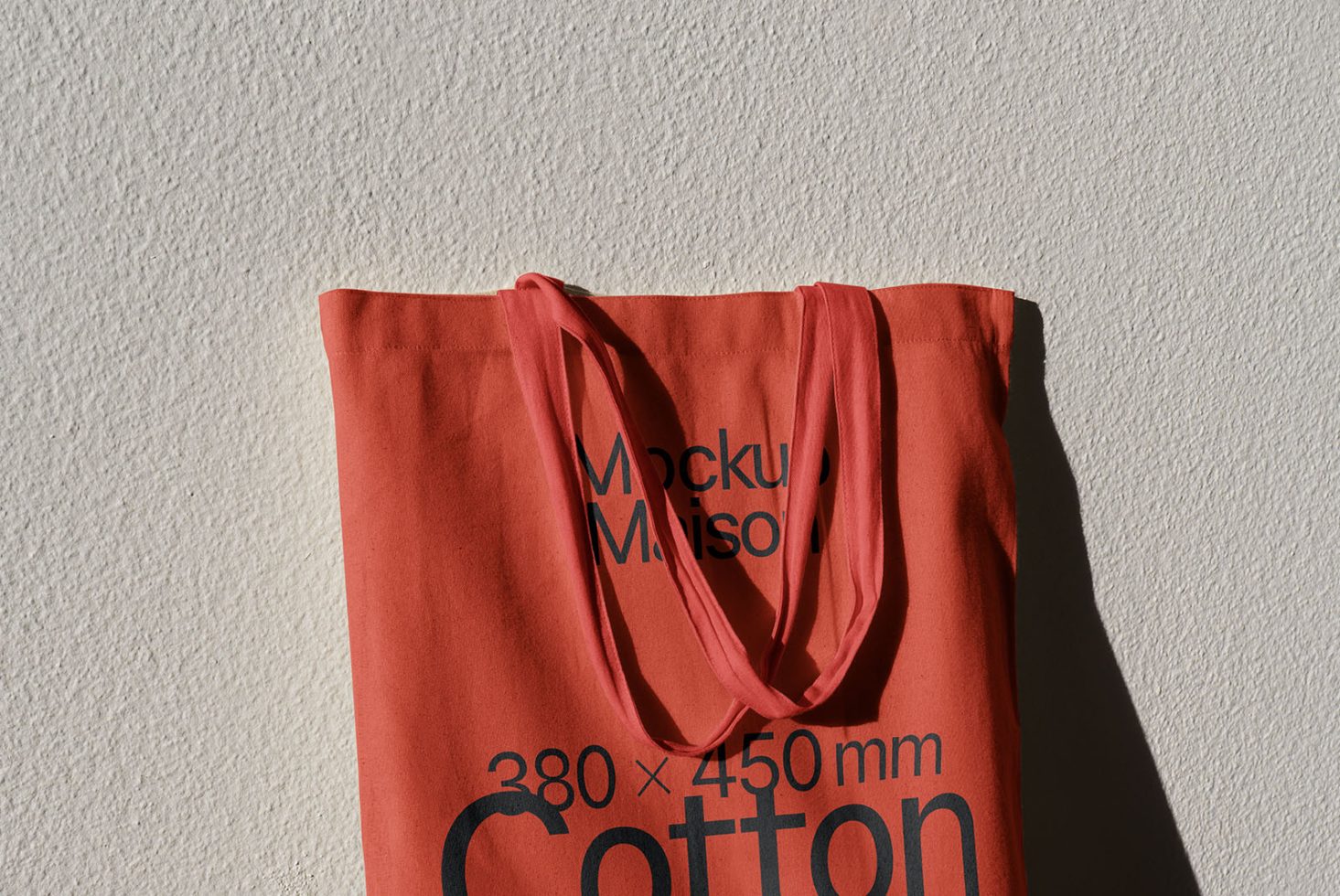 Red cotton tote bag mockup against textured wall, showing realistic shadows and fabric texture, ideal for showcasing design work.