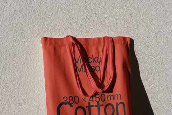 Red cotton tote bag mockup against textured wall, showing realistic shadows and fabric texture, ideal for showcasing design work.