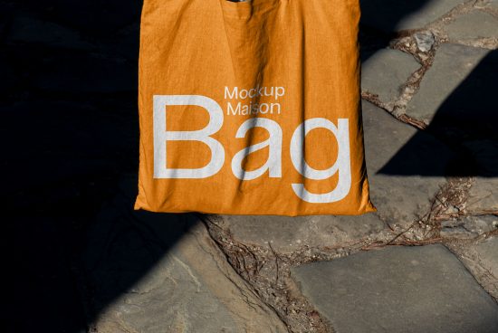 Orange tote bag mockup laying on stone pavement with bold typography design, realistic shadows, ideal for graphic presentations.