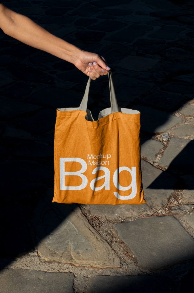 Person holding orange tote bag mockup with text design, outdoor setting, ideal for presentations in graphic design, templates, and mockups.