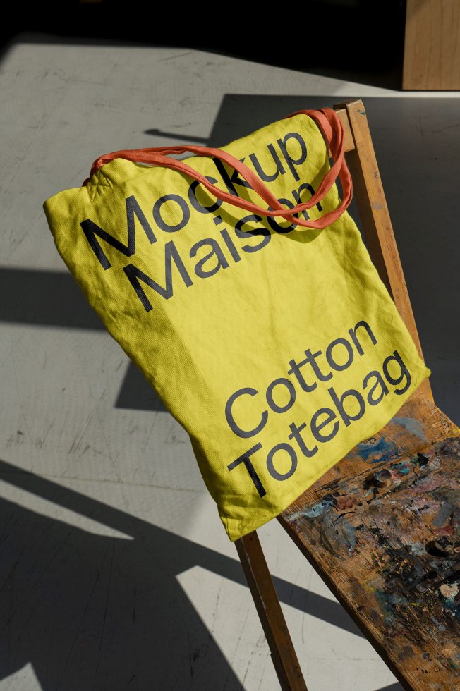 Yellow cotton tote bag mockup with black typography design on chair, natural lighting, designer asset for branding presentation.