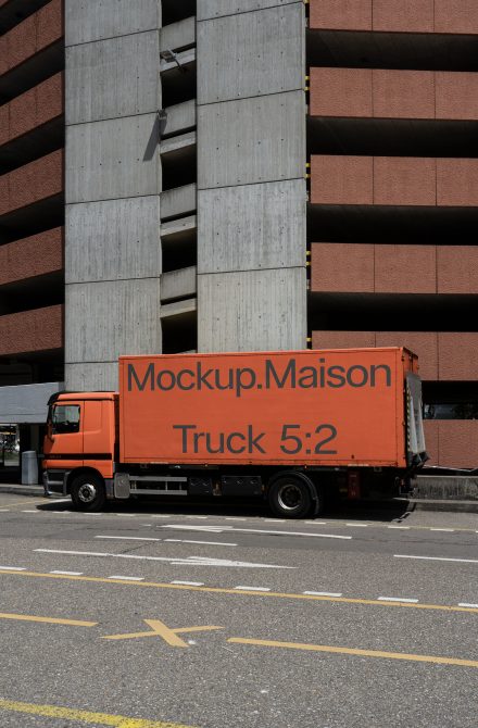 Orange delivery truck mockup for branding in urban setting with concrete building background, perfect for graphic design vehicle wrap visualization.