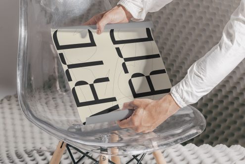 Person holding paper with bold typography design on clear chair, showcasing modern font style, essential for graphic design mockups.