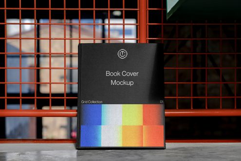 Black book cover mockup standing on ground against red metal grid fence