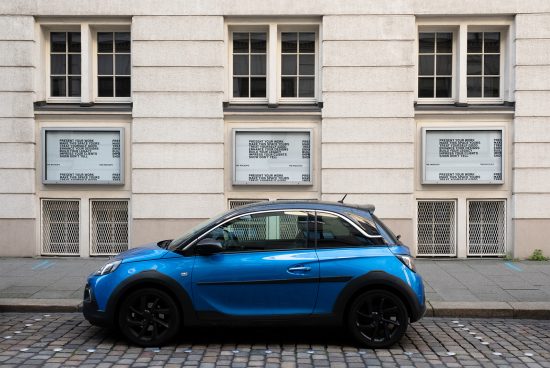 Mockup of blue compact car parked in urban setting with posters in building windows for showcasing design projects, ideal for vehicle branding.