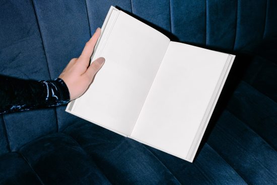 Person holding an open blank book for mockup on blue velvet sofa, ideal for presenting design templates and fonts.