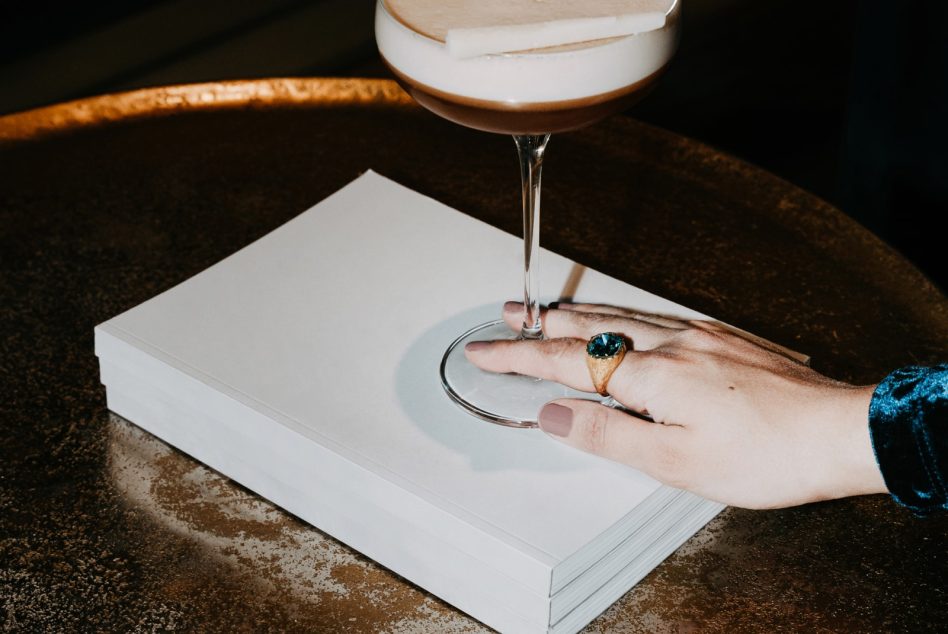 Hand holding cocktail glass over open notebook with jewelry on marble table