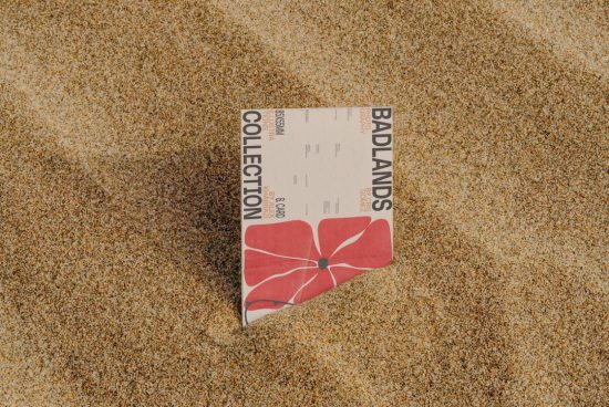 Mockup of a square booklet cover partially buried in sand, with a red flower design, ideal for presenting outdoor-related design work.