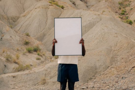 Person holding blank poster mockup in a desert landscape, ideal for design presentations and advertising mockups.