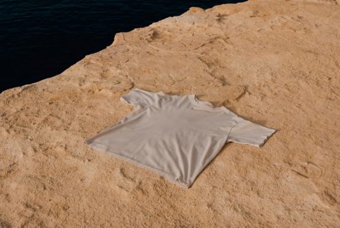 Plain t-shirt mockup on sandy rock by the sea, realistic apparel presentation, clothing design template, fashion graphic asset.