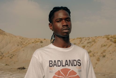 Serious young man in a white graphic t-shirt with 'Badlands' text, outdoor desert background, ideal for fashion mockups and portrait templates.