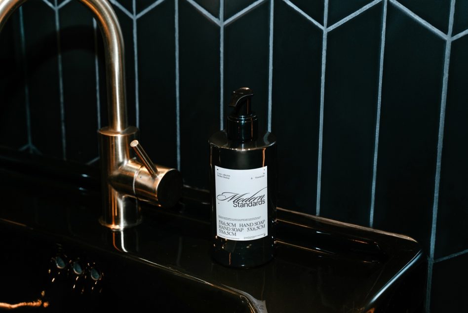 Black soap dispenser bottle with white label next to brass faucet on dark geometric tile
