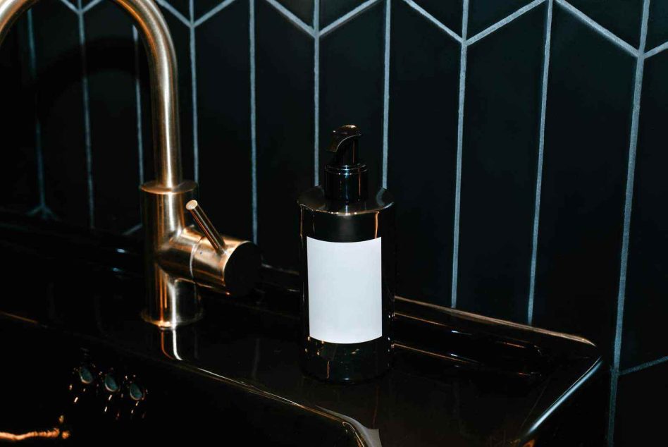 Black soap pump with blank white label next to gold tap on dark patterned tile wall