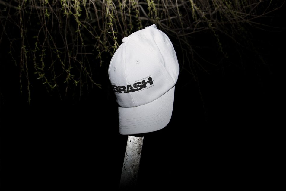 White baseball cap with dark BRASH text on front near hanging ivy backdrop