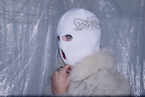 White knit ski mask with curved graffiti design on forehead, worn with white fur collar
