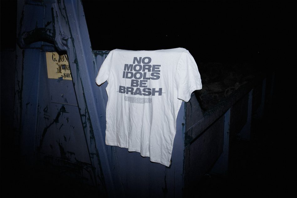 White shirt reading no more idols be brash on navy metal wall