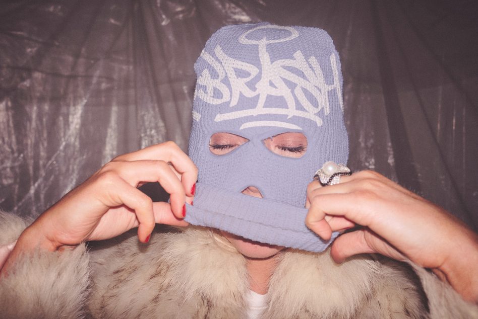 Person wearing lavender ski mask with graffiti lettering, hands adjusting balaclava