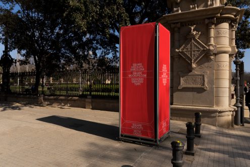 Red square kiosk display in Barcelona park by ornate stone column