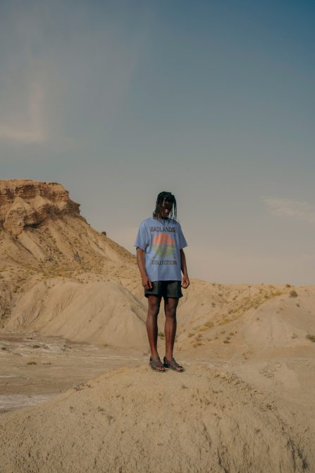 Person standing in desert landscape wearing graphic t-shirt, potential for fashion mockup template, with clear blue sky and textured sand.