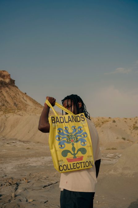 Person holding yellow tote bag with graphic design in desert landscape, potential mockup for showcasing bag designs, eco-friendly fashion.