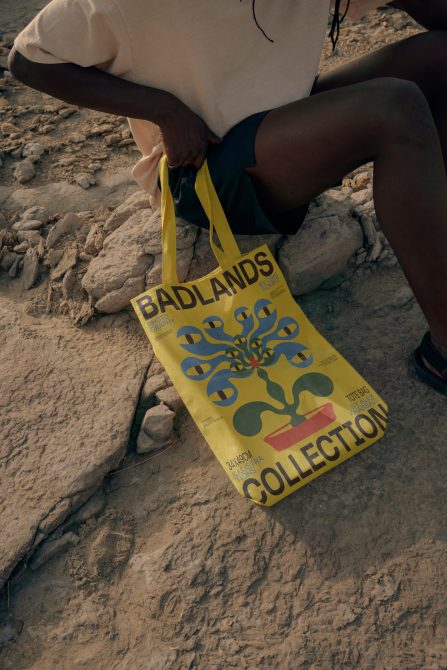 Yellow tote bag with Badlands Collection branding held by person sitting on sandy beach