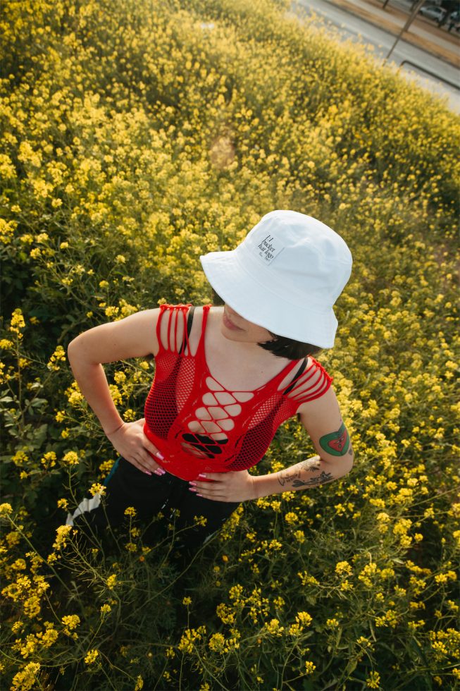 Woman in a red mesh top and white bucket hat posing in a yellow flower field, ideal for fashion mockups, outdoor apparel design.