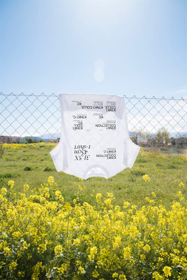 White t-shirt mockup hanging on a fence with flipped text design, outdoor spring setting with yellow flowers, clear sky for graphic display.