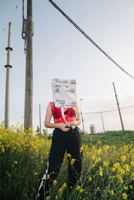 Person with bag over head standing in field of yellow flowers, urban setting, creative graphics for surreal design inspiration.