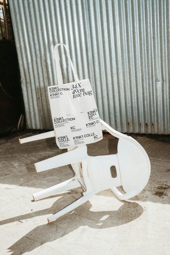 White tote bag with black text on it, hanging on an abstract wooden chair, industrial background, design mockup, branding presentation.