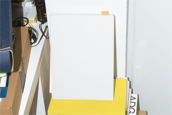 Blank vertical book cover mockup with a yellow bookmark on a cluttered workstation for design presentation.
