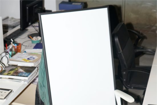 Blank white poster mockup on a black frame in an office setup with desk clutter, suitable for graphic design presentations.