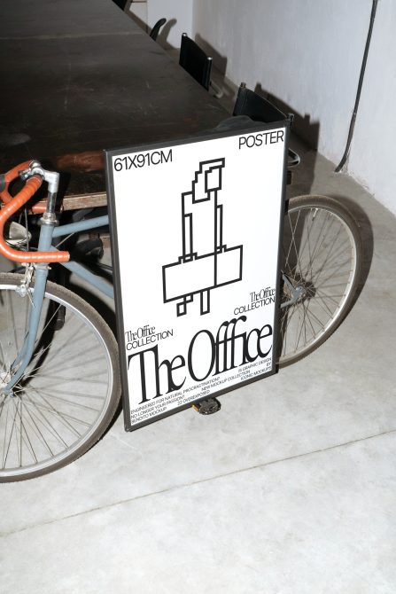 Poster mockup leaning on a bike against a textured wall in an urban setting, showcasing a black and white office-themed design.