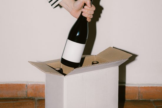 Hand placing a labeled wine bottle into cardboard box for mockup, ideal for designers to showcase branding and packaging designs.