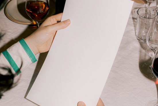 Person holding blank menu mockup at a dining table with elegant glassware, ideal for restaurant branding designs.