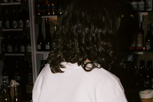 Person with curly hair facing bar shelves with bottles, moody ambiance for mockup or graphic design backgrounds.