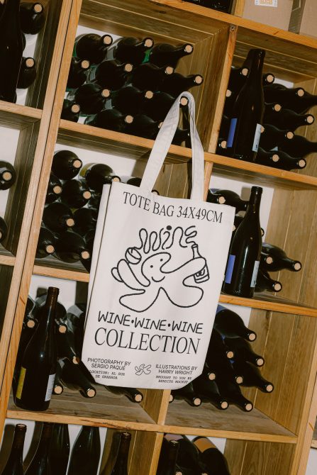 Canvas tote bag mockup with wine theme design hanging on wooden wine rack, ideal for showcasing bag designs and illustrations in a lifestyle setting.