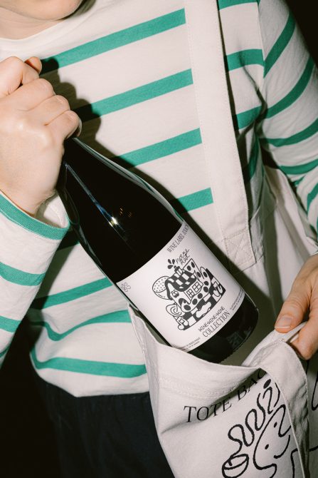 Person holding wine bottle with illustrated label, featuring graphic design elements, in front of striped shirt and tote bag.