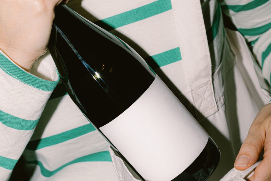 Person holding a wine bottle with blank label, ideal for mockup designs, branding, packaging, advertising, with striped shirt detail.