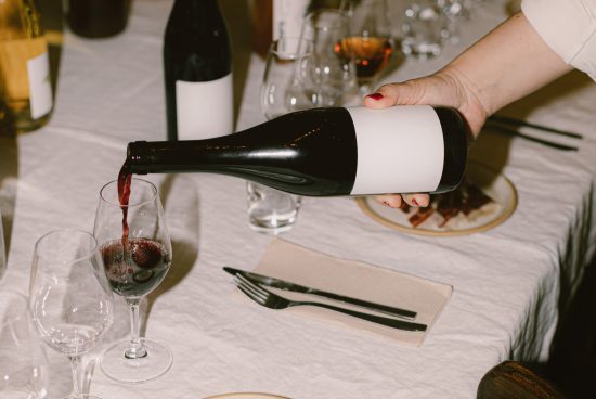 Hand pouring red wine into glass with empty label for design mockup, table setting background, food and drink concept.