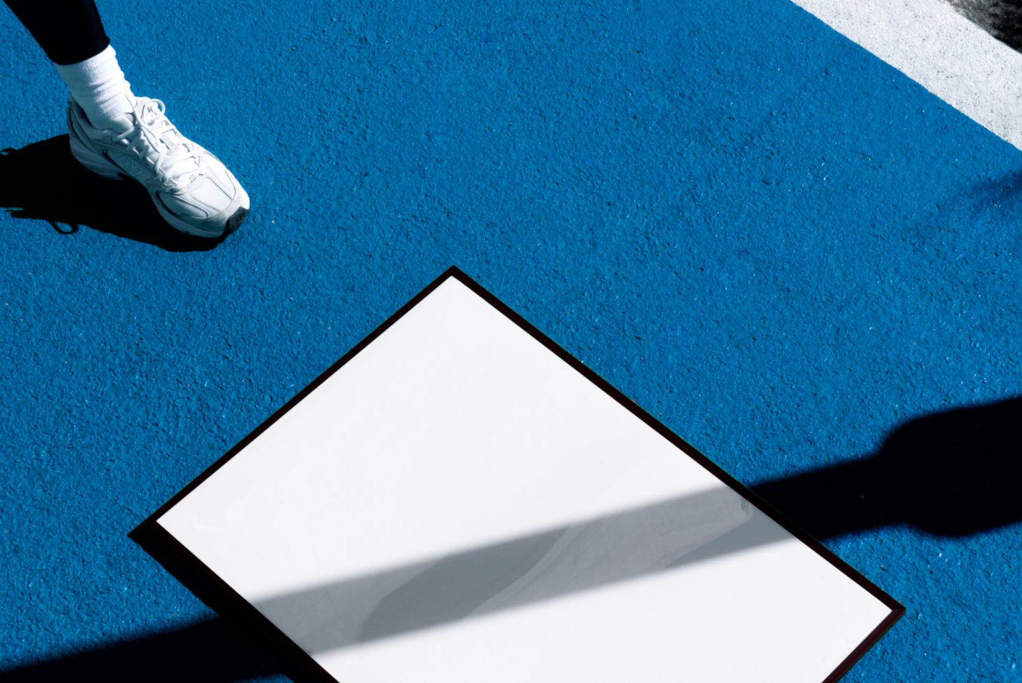 Person stepping near a white blank square poster mockup on a bright blue surface, with shadows casting, ideal for design presentations.