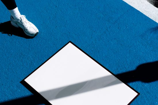 Person stepping near a white blank square poster mockup on a bright blue surface, with shadows casting, ideal for design presentations.