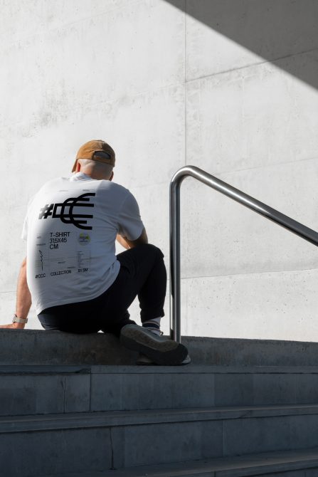 Man in white graphic t-shirt sitting on stairs for mockup display, modern urban clothing, concrete background, design template.