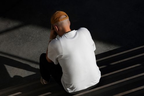 Man in blank white t-shirt and cap sitting on stairs for apparel mockup design, high-angle view, urban style, shadow pattern, fashion.