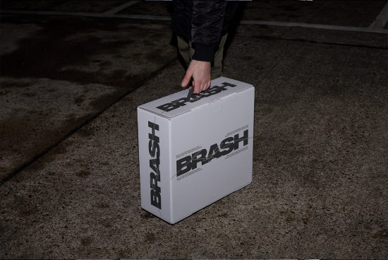 Person holding a branded cardboard box with bold typography design on urban background, ideal for mockup graphics and packaging design templates.