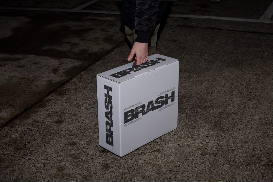 White cardboard box with BRASH branding held by hand on concrete ground